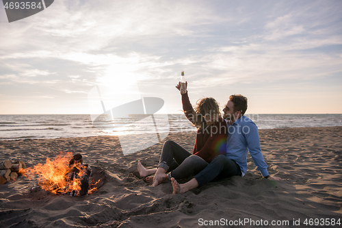 Image of Young Couple Sitting On The Beach beside Campfire drinking beer