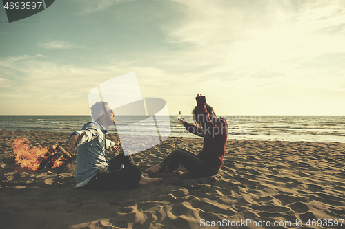 Image of Young Couple Sitting On The Beach beside Campfire drinking beer