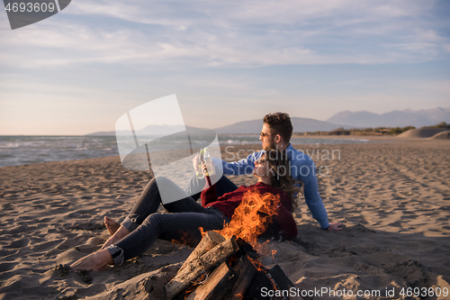 Image of Young Couple Sitting On The Beach beside Campfire drinking beer