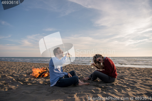 Image of Young Couple Sitting On The Beach beside Campfire drinking beer