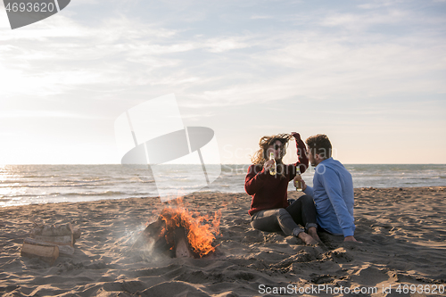 Image of Young Couple Sitting On The Beach beside Campfire drinking beer