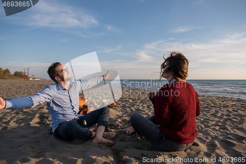 Image of Young Couple Sitting On The Beach beside Campfire drinking beer