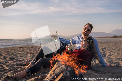 Image of Young Couple Sitting On The Beach beside Campfire drinking beer