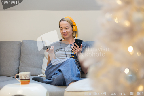 Image of Young cheerful woman sitting indoors at home living room sofa watching TV, using social media on phone for video chatting and staying connected with her loved ones