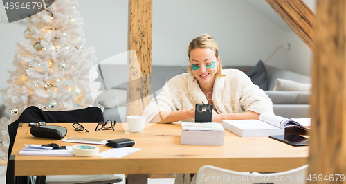 Image of Young businesswoman wearing cosy warm bathrobe and cosmetic eye patches working remotly from home in winter Christmas time during 2020 corona virus pandemic. Work from home, selter in place, concept