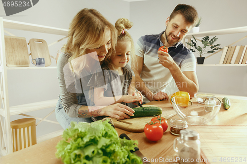 Image of Cute little girl and her beautiful parents are cutting vegetables in kitchen at home