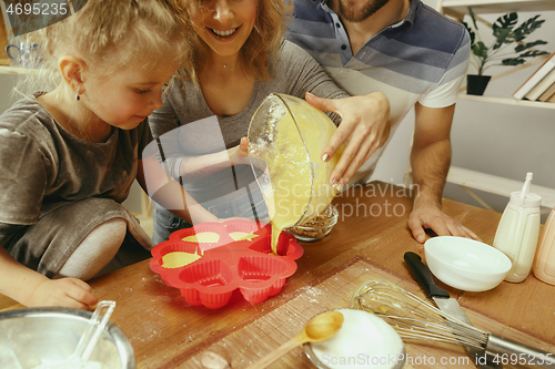 Image of Cute little girl and her beautiful parents preparing the dough for the cake in kitchen at home