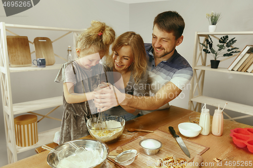 Image of Cute little girl and her beautiful parents preparing the dough for the cake in kitchen at home