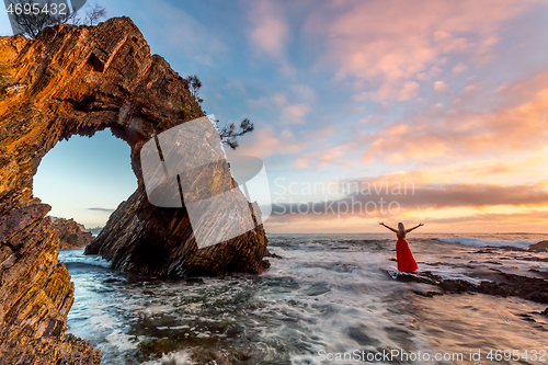 Image of Woman in long red dress standing in ocean waves by a sea arch