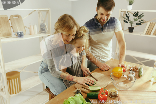 Image of Cute little girl and her beautiful parents are cutting vegetables in kitchen at home