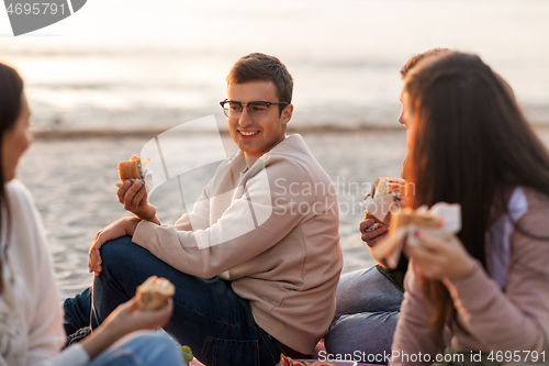 Image of happy friends eating sandwiches at picnic on beach