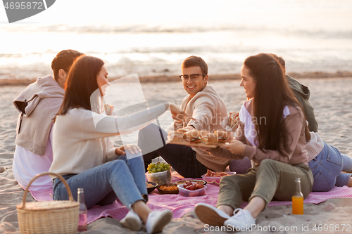 Image of happy friends eating sandwiches at picnic on beach