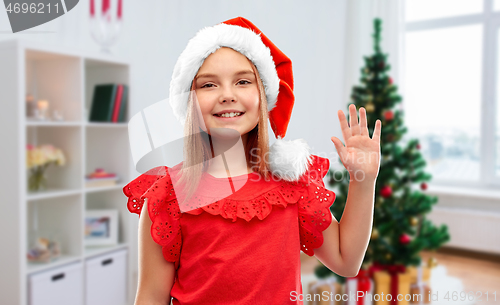 Image of happy girl in santa helper hat waving hand at home