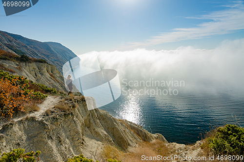 Image of A girl is standing on a rock who watches the clouds above the sea