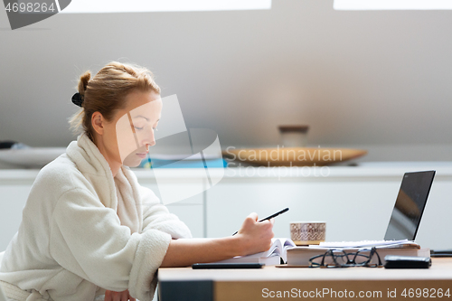 Image of Young female entrepreneur wearing cosy warm bathrobe working remotly from home in winter Christmas time during 2020 corona virus pandemic. Work from home, selter in place, concept.