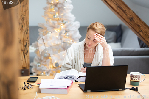 Image of Young female entrepreneur wearing cosy warm bathrobe working remotly from home in winter Christmas time during 2020 corona virus pandemic. Work from home, selter in place, concept.