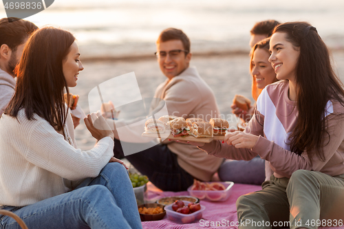 Image of happy friends eating sandwiches at picnic on beach