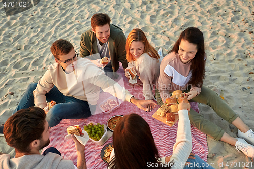 Image of happy friends eating sandwiches at picnic on beach