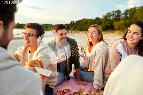 Image of happy friends eating sandwiches at picnic on beach