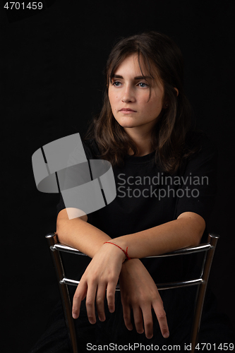 Image of Portrait of a young girl sitting on a high chair and folded her hands on the back of a chair, black background