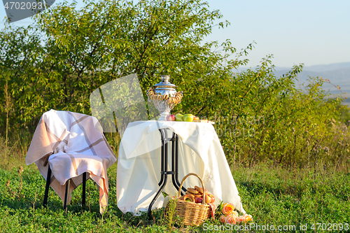 Image of On the field under the bushes is a table with a samovar and fruit, next to it is a chair