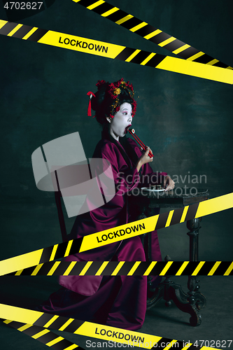 Image of Young japanese woman as geisha isolated on dark green background. Drinking cola and eating popcorn behind limiting message lockdown on the screen