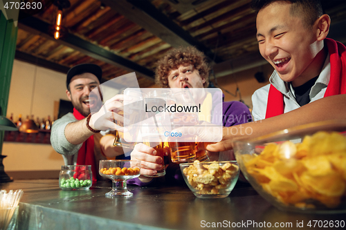 Image of Excited, shocked soccer fans cheering for favourite sport team in restaurant or bar behind the message with word Lockdown