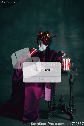 Image of Young japanese woman as geisha isolated on dark green background. Drinking cola and eating popcorn behind limiting message lockdown on the screen