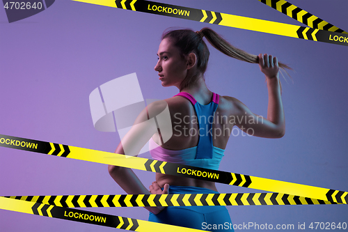 Image of Young sportive woman posing on gradient neon background behind limiting tapes with word Lockdown