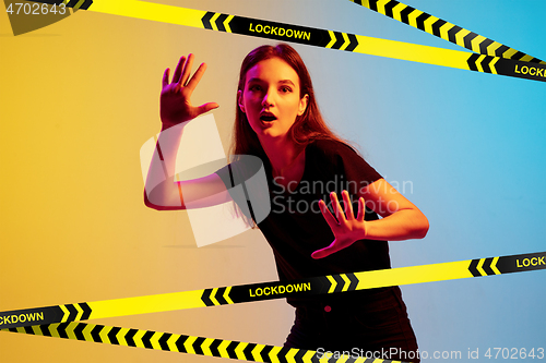 Image of Young sportive woman posing on gradient neon background behind limiting tapes with word Lockdown