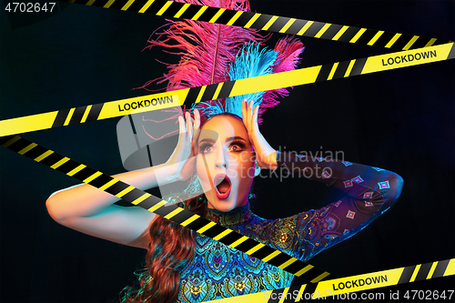 Image of Beautiful young woman in carnival, stylish masquerade costume with feathers behind limiting tapes with word Lockdown