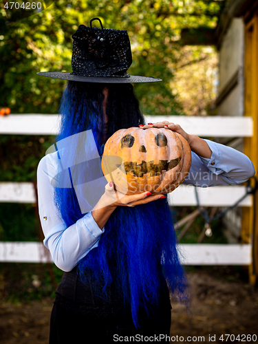 Image of A girl with her hair covered face in a witch costume is holding a pumpkin with a painted face and a witch\'s cap