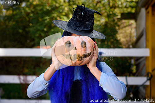 Image of A girl dressed as a witch covered her face with a pumpkin with an evil face drawn in the style of a Halloween celebration
