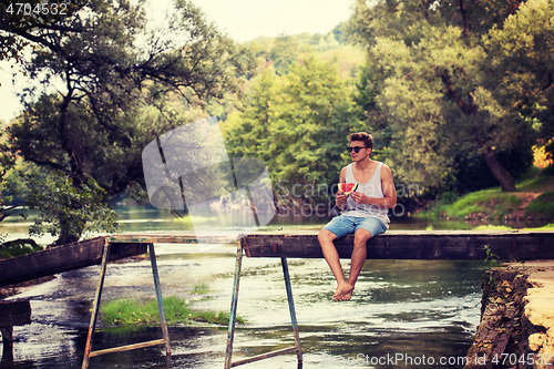 Image of man enjoying watermelon while sitting on the wooden bridge