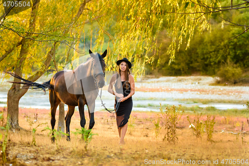 Image of A girl in a beautiful black dress and a black hat walks with a horse across the field