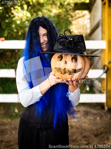 Image of A girl dressed as a witch with blue hair is holding a pumpkin with a painted face and a witch\'s cap