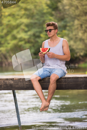 Image of man enjoying watermelon while sitting on the wooden bridge
