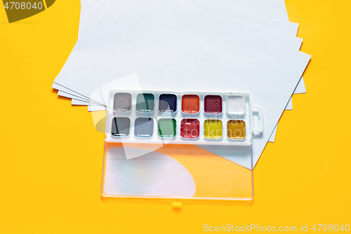 Image of Blank white sheets and 12 colors of watercolor paint on a yellow background