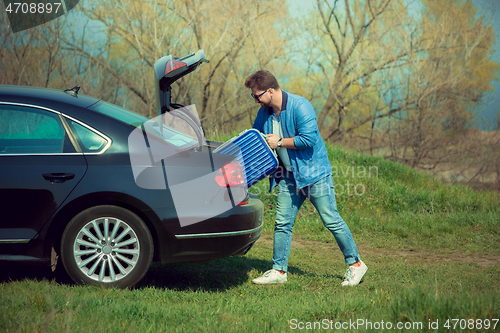 Image of Handsome smiling man going to vacations, loading his suitcase in car trunk