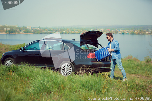 Image of Handsome smiling man going to vacations, loading his suitcase in car trunk