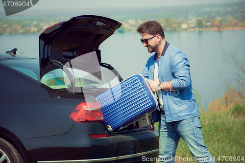 Image of Handsome smiling man going to vacations, loading his suitcase in car trunk