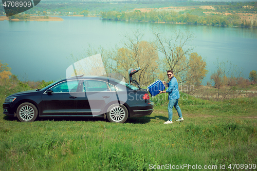 Image of Handsome smiling man going to vacations, loading his suitcase in car trunk