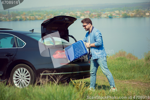 Image of Handsome smiling man going to vacations, loading his suitcase in car trunk