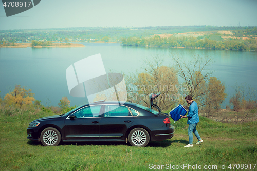 Image of Handsome smiling man going to vacations, loading his suitcase in car trunk