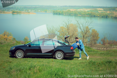 Image of Handsome smiling man going to vacations, loading his suitcase in car trunk