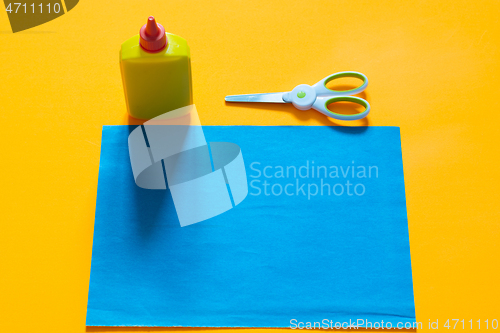 Image of On a yellow background lies a sheet of blue colored paper, next to it there is a tube of glue and scissors