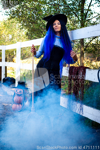 Image of A girl dressed as a witch stands at a fence on a farm, smoke from below, pumpkins in the background