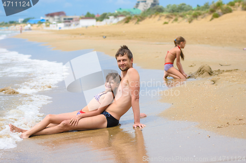 Image of Dad and daughter are sitting in the foreground, in the background a girl is building a sand castle