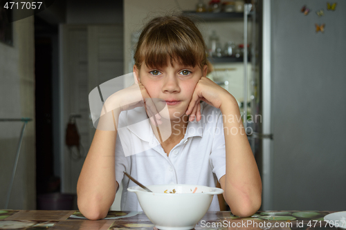 Image of The girl is sitting at the table in the kitchen, in front of her is an empty plate, the girl looks into the frame