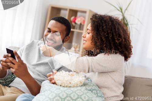 Image of african couple with popcorn and smartphone at home